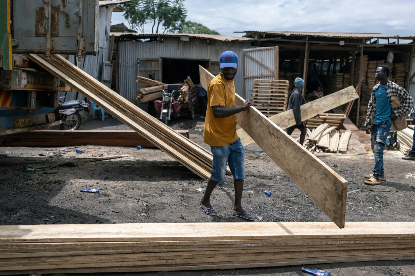 A man in a yellow T-shirt and denim shorts carries a long wooden plank in a lumber yard, amid stacks of timber and other people.