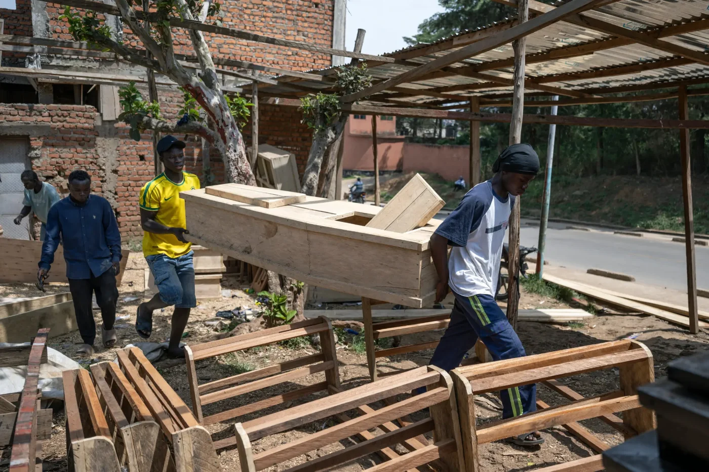 Four people are in a dusty outdoor area with wooden structures and brick buildings. Two of them carry a light-colored wooden coffin.