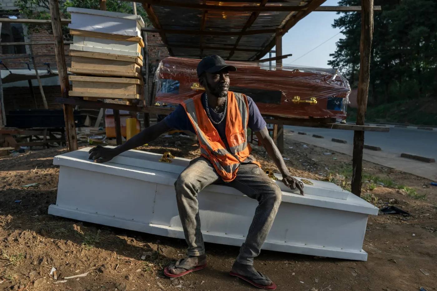 Mr. Limayo sits on a white coffin, wearing an orange vest and dark cap. Multiple coffins are displayed in an open-air workshop.